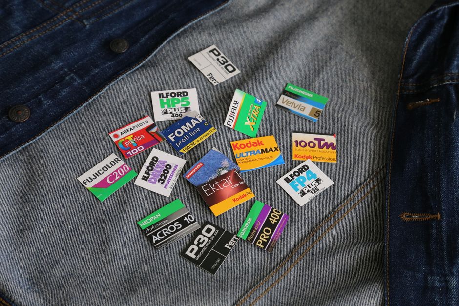 A close-up image showing a collection of various photographic film labels and packaging placed on a pair of blue denim jeans. The labels are made of paper and plastic, featuring bright colors, printed text, and brand logos, including well-known names like Kodak, Fuji, and Ilford. Some labels display technical details such as film type, ISO speed ratings, and product descriptions, with a mix of horizontal and vertical orientations. The jeans have a textured, slightly faded finish with visible stitching and metallic rivets at the pocket corners, illustrating casual wear. The background appears to be a neutral surface, possibly a fabric or environment where the jeans are laid flat, with natural lighting that highlights the vivid colors and textures of both the labels and denim. This scene aligns with the concept of individual disposal or organization of photographic materials, subtly tying into alternative waste handling for specialized items by private waste removal services such as Waste Removal Ilford, who support various waste collection needs including niche or technical waste streams.