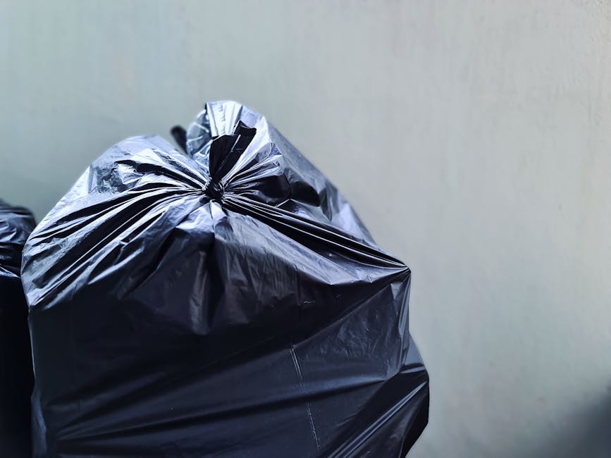 Close-up of three black plastic rubbish bags filled with waste, tightly tied at the top, placed against a plain light grey wall in an outdoor setting. The bags appear to be made of glossy, durable material with noticeable creases and folds, indicating they are full and sealed for collection. The foremost bag is positioned slightly to the left, with two additional bags partially visible behind it, stacked or leaning against each other. The scene suggests a private waste collection setup, possibly prepared for on-site clearance or alternative disposal separate from regular municipal rubbish removal. The neutral lighting highlights the shiny surface of the bags, emphasizing their contents and texture. This image is relevant to professional waste management services like those provided by Waste Removal Ilford, who handle private rubbish collections and on-demand waste removal needs in the local area.