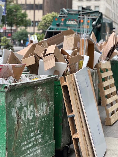 A sanitation worker dressed in a blue uniform and orange high-visibility vest is emptying a blue waste bin into the back of a white refuse collection truck parked on a narrow street. The truck is equipped with a hydraulic lifting mechanism at the rear, showing signs of rust and wear on the metal surface. The street is lined with multi-story residential buildings constructed from light-colored brick and concrete, with some areas displaying patches of weathered paint or plaster. A small, black vehicle with visible license plates is parked on the left side of the street, and two road signs indicating parking restrictions and no entry are attached to a pole on the sidewalk. The environment appears overcast, with soft natural lighting illuminating the scene, which is typical for an urban area where private rubbish collection services might be employed as an alternative to council-led waste disposal, aligning with the services offered by Waste Removal Ilford.
