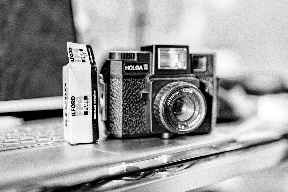A black-and-white photograph depicts a vintage camera with a textured, black body and a prominent, ridged lens, positioned on a flat surface, likely a desk or table. To the left of the camera, there is a small box of Ilford Plus black and white photographic film, which is upright and slightly taller than the camera. The film box displays bold text and branding, with the label indicating it is for black-and-white photography. The background is out of focus but suggests an indoor environment, possibly a workspace or studio, with objects faintly visible behind the camera and film box. The lighting appears soft, highlighting the textured surface of the camera and the glossy finish of the film packaging without harsh shadows. The scene presents a neat arrangement of photographic equipment, potentially for use in a private collection or photo-laboratory setting, aligning with services related to private waste handling or collection of personal or professional artifacts. Waste Removal Ilford’s presence could be inferred in the context of clearing or disposing of older photographic materials or equipment through independent collection services, reflecting a practical approach to managed waste removal of delicate or specialized items.