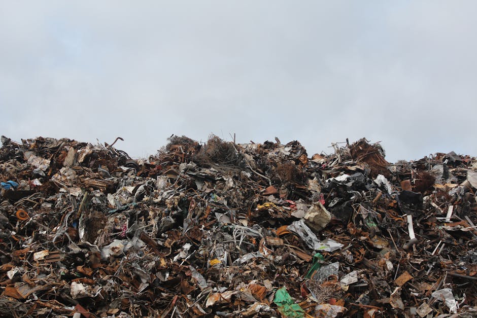 A large pile of mixed industrial and household waste materials extends across the foreground, consisting of various types of debris including rusted metal parts, broken plastic fragments, crumpled cardboard, and scattered, shredded paper. The materials display different textures such as jagged metal edges, smooth plastic surfaces, and coarse cardboard, with colors ranging from metallic rust tones and dark plastics to lighter browns and greens. The pile is densely packed, with objects layered and intertwined, forming a mound that rises towards the sky. In the background, there is a cloudy, overcast sky providing diffuse lighting that highlights the irregular surfaces and diverse textures of the refuse. The scene appears to be outdoors on a flat surface, possibly a landfill or an unmanaged dumping site, illustrating the type of waste that companies like Waste Removal Ilford might help manage through private or alternative rubbish collection methods, reducing illegal dumping in urban areas.