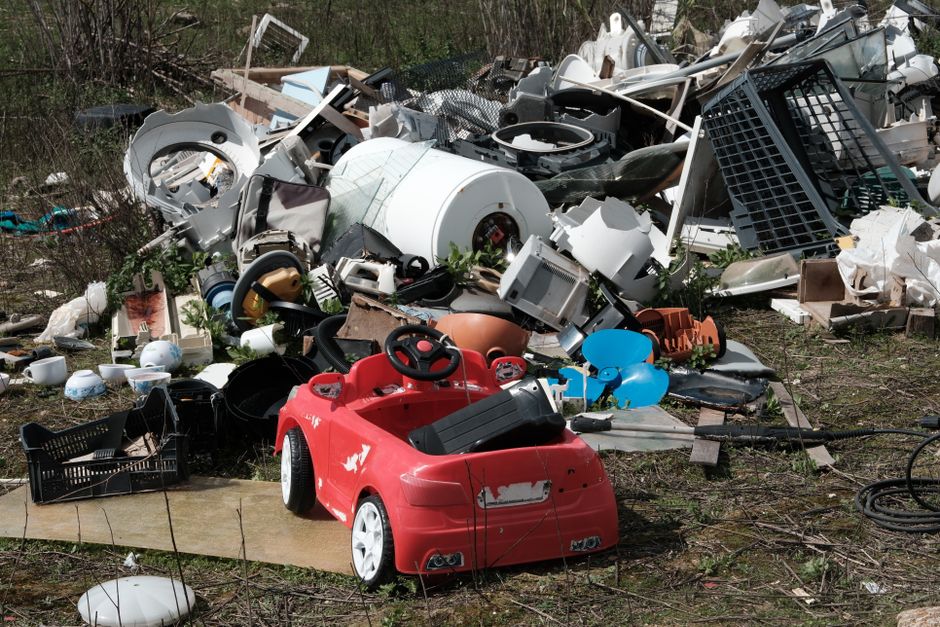 A large pile of discarded household waste and debris is situated outdoors, consisting of various items including a small red plastic child's ride-on car positioned in the foreground with minor scratches and paint chips, and other objects such as a white washing machine drum, black plastic crates, broken ceramic dishes, soft furnishings, and fragments of furniture. The waste is spread across an uneven grassy and dirt area, with some items leaning on each other and others scattered on the ground. In the background, overgrown vegetation and trees can be seen, indicating an unmanaged, possibly neglected outdoor space. The lighting suggests natural daylight, casting soft shadows on the cluttered scene. This scene relates to private waste disposal or illegal dumping, highlighting the importance of proper rubbish removal services to handle such clutter, as provided by Waste Removal Ilford, ensuring environmentally responsible clearance of household and garden waste.
