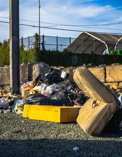 A pile of mixed waste and rubbish bags, primarily black plastic, is situated on a gravel surface next to a low stone wall in an outdoor area. Among the debris, there is a large, yellow plastic container lying on its side and an old, worn beige car seat with fabric upholstery leaning against the rubbish. Behind the pile, a tall metal pole supports overhead electrical or telephone wires that stretch across the sky. In the background, a dense hedge borders the scene, with a tennis court or sports facility enclosed by a mesh fence and a large, curved, partially transparent canopy structure. The scene is lit by natural daylight, with clear blue skies and some wispy clouds, indicating a sunny day. The arrangement of waste and discarded furniture suggests an instance of private rubbish disposal or on-site clearance, reflecting the service scope of waste removal providers like Waste Removal Ilford.