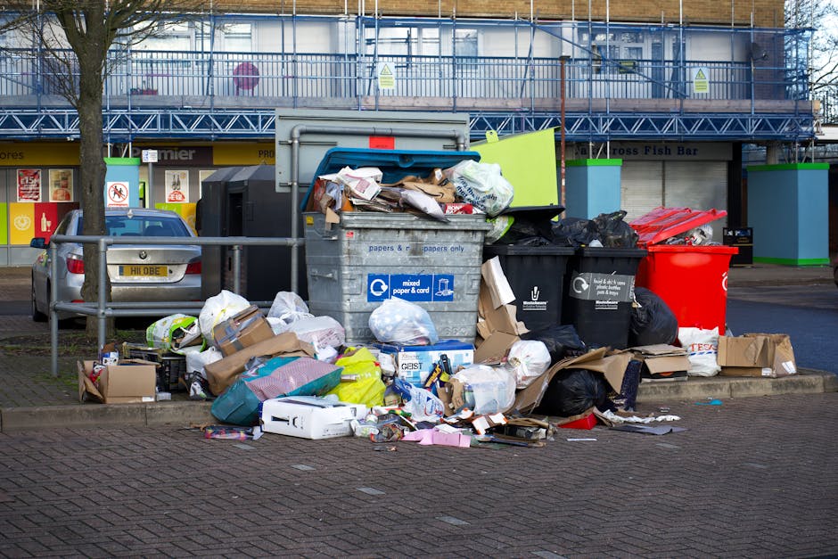 A collection of waste and rubbish accumulated on a paved urban sidewalk, featuring multiple black, black and red, and grey plastic wheelie bins overflowing with various types of discarded materials such as cardboard boxes, paper waste, plastic bags, and packaging. The waste extends onto the ground, with torn and crumpled cardboard, plastic wrappers, and loose paper scattered in front of the bins. One large grey bin has a partially open lid revealing mixed paper and cardboard inside, while black and red bins next to it contain black plastic bags and other refuse. Behind the rubbish, a metal railing separates the area from a parking lot where a silver car is parked. In the background, a building with storefronts and signage is visible, with a blue scaffolding structure covering part of the upper facade. The overall scene suggests an area awaiting waste collection, aligned with private waste handling services such as those offered by Waste Removal Ilford, illustrating the need for efficient rubbish removal and site clearance.