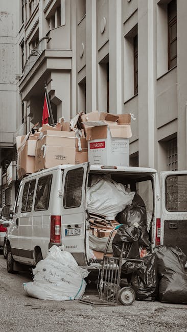 A white van parked on a city street with its rear doors open, revealing a large pile of mixed waste materials including black and white plastic rubbish bags, cardboard boxes, and loose packaging materials. Several cardboard boxes, some torn and flattened, are stacked on top of the van, while others lean against its roof. A shopping trolley is positioned next to the open tailgate, filled with additional black plastic bags. The van is situated beside a multi-storey building with a grey stucco exterior, featuring rectangular windows and decorative architectural details in a neutral colour palette. The scene suggests an instance of private waste collection or on-site clearance, with waste materials temporarily stored for removal by a professional rubbish removal service such as Waste Removal Ilford. The lighting appears subdued, indicating an overcast day, and the environment is typical of an urban setting where immediate rubbish disposal is being managed effectively outside the premises, supporting alternative waste handling outside of local authority collection methods.