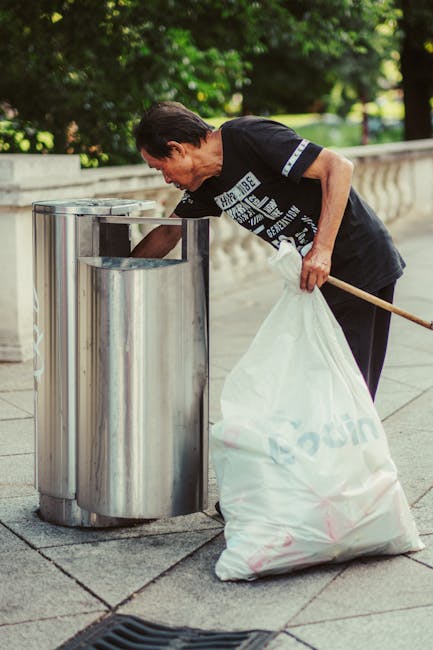 A middle-aged man with short dark hair, wearing a black T-shirt with white text and dark pants, is seen bending over while disposing of a white plastic bag filled with waste into a stainless steel litter bin on a paved outdoor walkway. The bin has a modern, polished finish with a rectangular opening on its top, situated against a backdrop of lush green trees and bushes, suggesting a park or garden setting. The man holds a wooden stick in his right hand, used to lift or open the bin’s lid, which is partially retracted to accommodate the bag. The surrounding environment is well-maintained, with a stone balustrade or railing visible to the right, and natural sunlight illuminating the scene, highlighting the reflective surface of the bin and the greenery behind. This scene exemplifies private waste disposal practices in a public outdoor space, underscoring the role of rubbish removal services and independent waste collection in maintaining cleanliness and environmental standards.