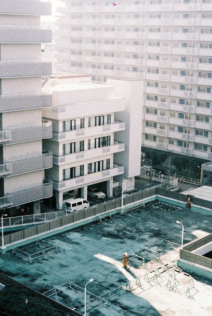 The image shows a rooftop area of a multi-storey residential building with a flat, asphalt surface that appears weathered and slightly worn. The rooftop is surrounded by a low metal railing, with several bike racks installed along the edges, some of which are empty. A few small, orange utility pipes and vents are visible protruding from the surface, along with a white street lamp at the bottom right corner. In the background, there are tall, uniform apartment blocks with numerous windows and balconies, primarily constructed with concrete and painted in light shades of grey or off-white. On the ground level beneath the rooftop, parked cars are visible within a designated parking area, partially sheltered by supporting columns of the building. The surrounding environment suggests an urban setting, with dense high-rise buildings affecting the natural light and casting shadows across the rooftop. The scene is illuminated by natural daylight, with soft, diffuse lighting indicating a partly cloudy day. The setting appears to be part of a managed residential complex, possibly involving a shared outdoor space, and aligns with the concept of private property management related to rubbish and waste handling through external services like Waste Removal Ilford, which may facilitate on-site clearance or bulk waste collection for communal areas.