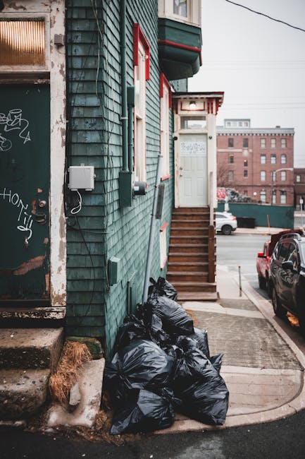 The image depicts a narrow urban alleyway adjacent to a building with green shingle siding and red-trimmed windows, leading up to a set of wooden stairs with a white door at the top. Several black rubbish bags are piled on the concrete pavement at the building's base, positioned on the left side near a weathered, graffiti-marked metal door with peeling paint, and a small patch of litter behind the bags. Behind the alley, parked cars are visible along the street, with a large brick building in the background under an overcast sky. The scene suggests a typical residential or mixed-use area where waste collection may be handled privately, possibly by a waste removal service like Waste Removal Ilford, to manage refuse outside properties where access for council rubbish collection may be restricted or unavailable. The overall environment appears clean but cramped, with the bags likely awaiting collection or removal, emphasizing the need for efficient rubbish disposal solutions in urban settings.